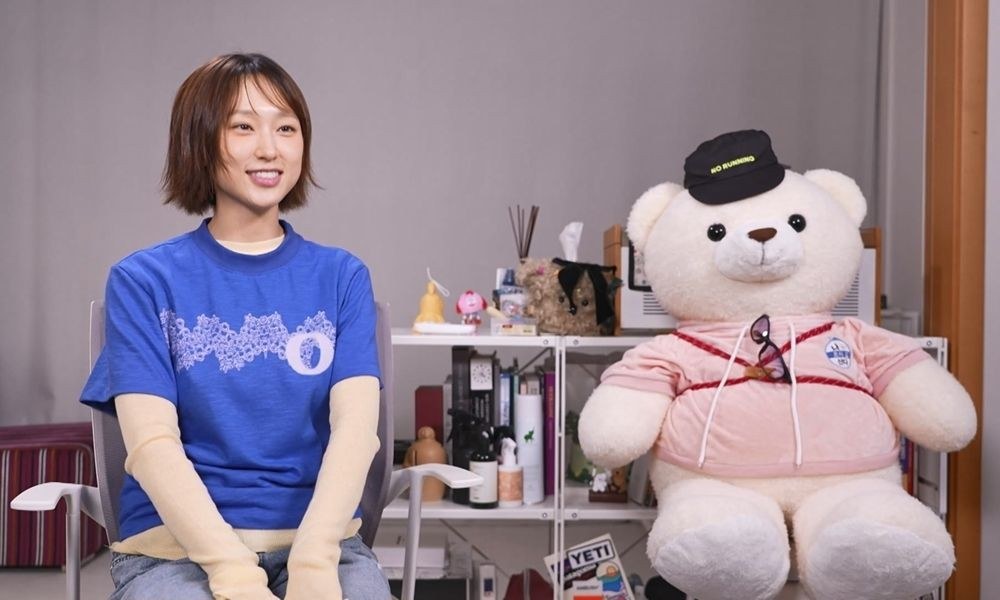 선반 위 소품과 대형 곰인형을 배경으로 앉아 인터뷰하는 류혜영