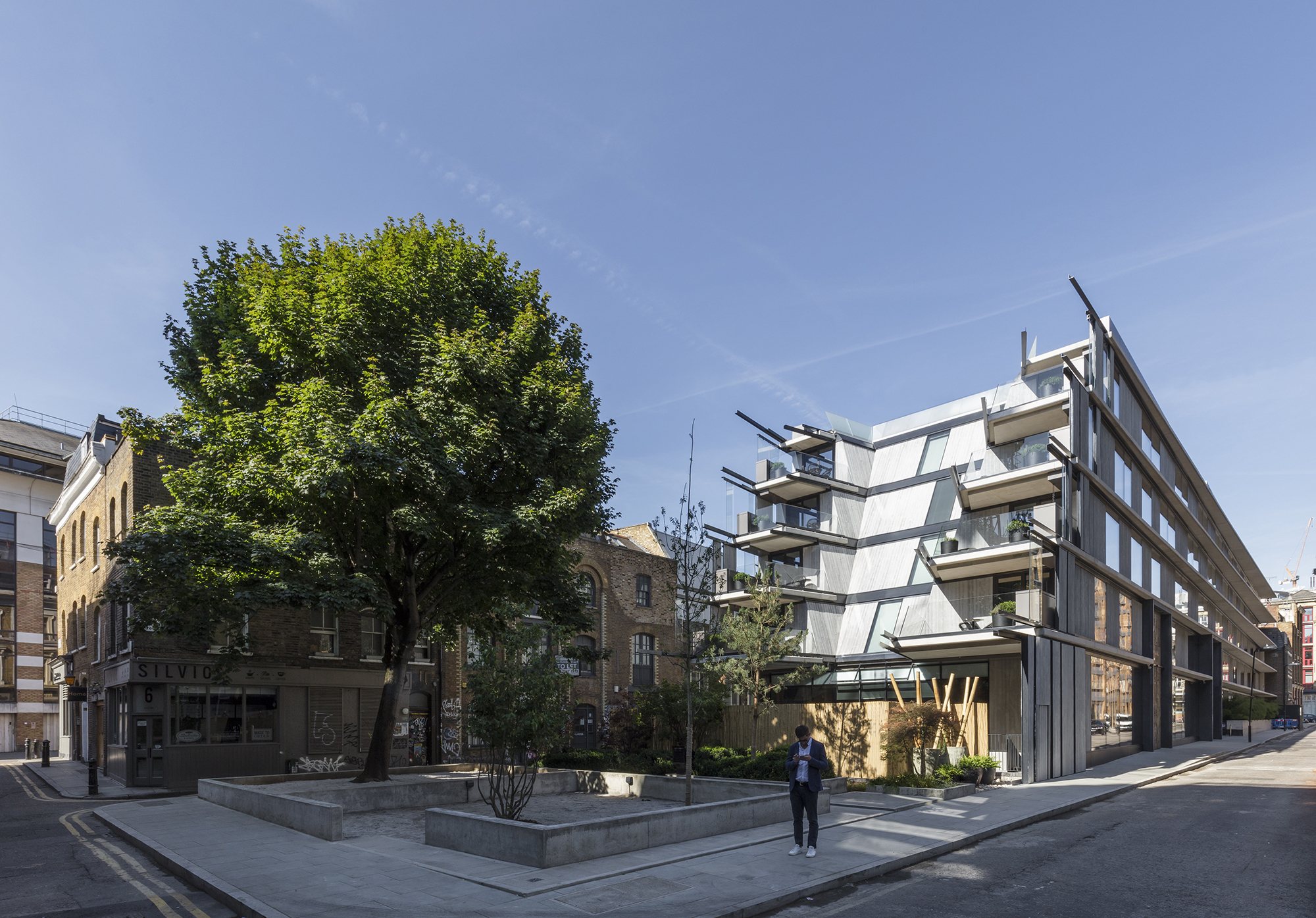 쇼디치 Willow Street 모퉁이 광장과 나무 너머로 보이는 Aethos Shoreditch 건물 외관